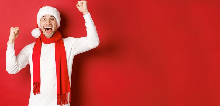 Image of joyful caucasian man in santa hat and scarf, shouting for joy and raising hands, celebrating victory or win, triumphing over red background.