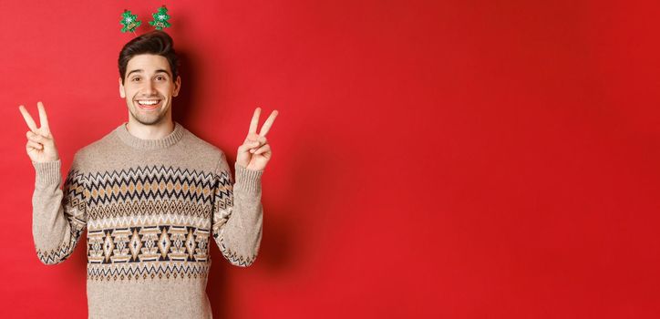Concept of winter holidays, christmas and celebration. Image of handsome and silly guy dressed for new year party, showing peace signs and smiling, standing against red background.