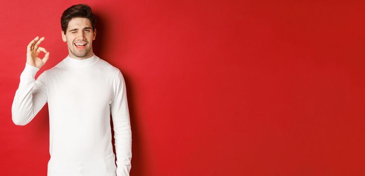 Portrait of charismatic beaded guy in white sweater, assure you, winking and showing okay sign to guarantee good quality, praise excellent choice, standing over red background.