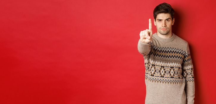 Image of serious handsome man extending one finger to stop you, telling no, give warning, standing over red background in winter sweater.