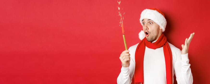 Concept of christmas, winter holidays and celebration. Close-up of amazed handsome man in santa hat and scarf, looking at sparkler with excitement, standing over red background.