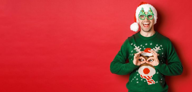 Portrait of carefree handsome man in santa hat and party glasses, making fun of his christmas sweater, looking happy over red background.