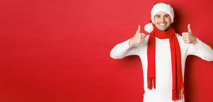 Portrait of handsome smiling man in santa scarf and hat, showing thumbs-up, celebrating christmas, standing over red background.
