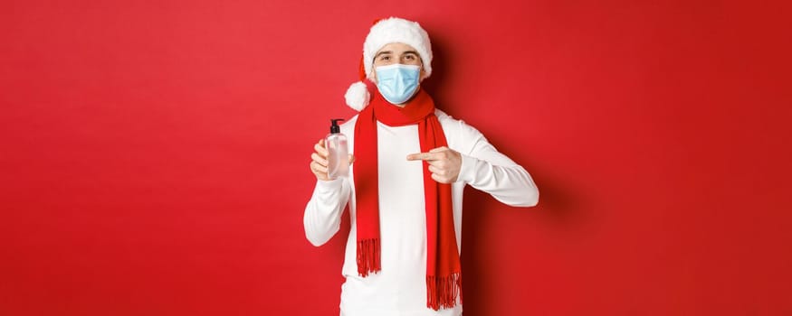 Concept of covid-19, christmas and holidays during pandemic. Handsome happy man in santa hat and medical mask, recommending use hand sanitizer, standing over red background.