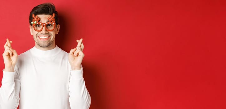 Close-up of excited handsome man, wearing party glasses and white sweater, crossing fingers for good luck, celebrating christmas and waiting wish, anticipating gifts, standing over red background.
