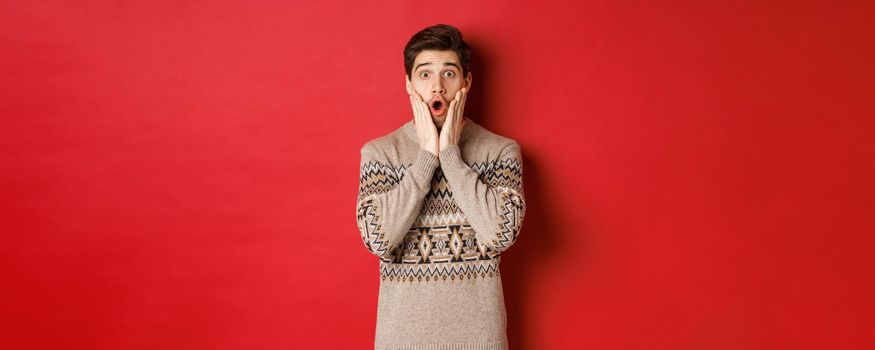Image of surprised handsome guy reacting to cool new year promo offer, gasping amazed, wearing christmas sweater, standing over red background.
