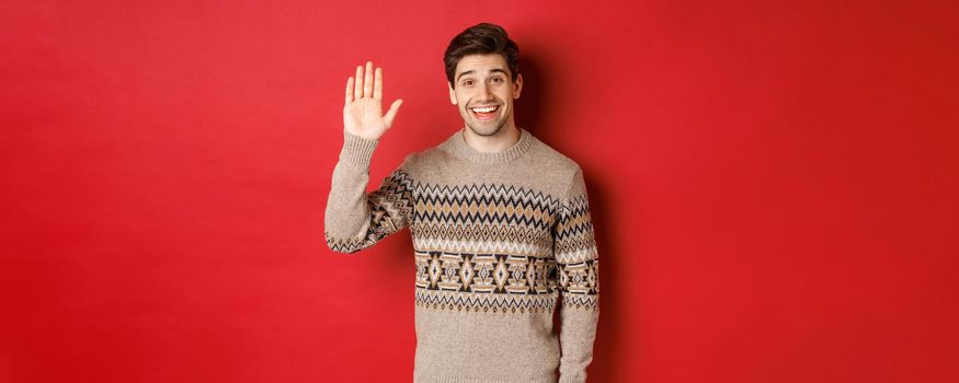 Portrait of handsome and friendly guy in christmas sweater, saying hello and waving hand, greeting someone and wishing happy winter holidays, standing over red background.