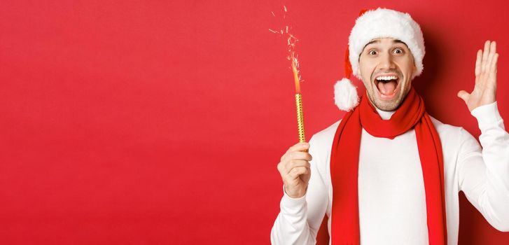 Concept of christmas, winter holidays and celebration. Handsome man celebrating new year and having fun, holding sparkler and smiling, wearing santa hat, standing over red background.