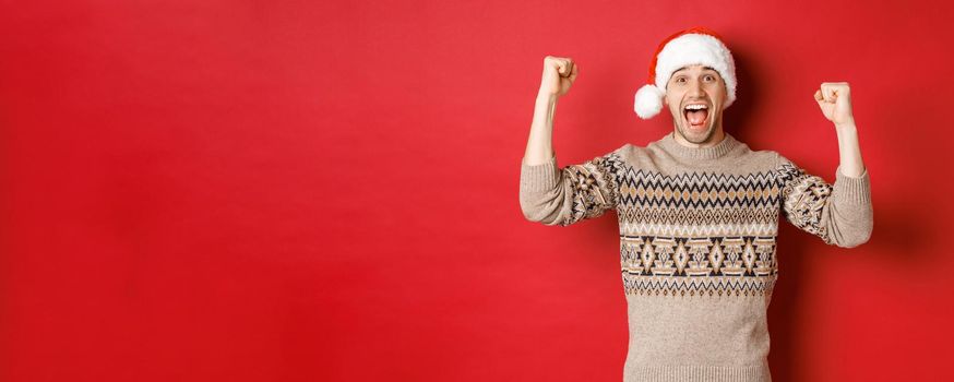 Image of cheerful handsome man in swearer and santa hat, celebrating new year, triumphing or winning something, raising hands up and shouting for joy, standing over red background.