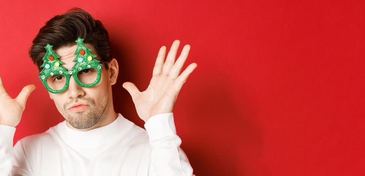 Concept of winter holidays, christmas and celebration. Close-up of funny handsome guy in party glasses, showing empty hands, standing over red background.