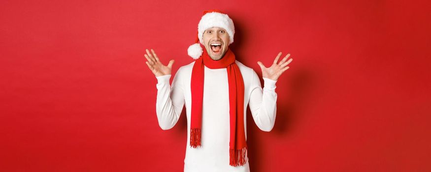 Portrait of happy and amazed handsome man, celebrating new year, wishing merry christmas, wearing santa hat and scarf, telling big news, standing against red background.
