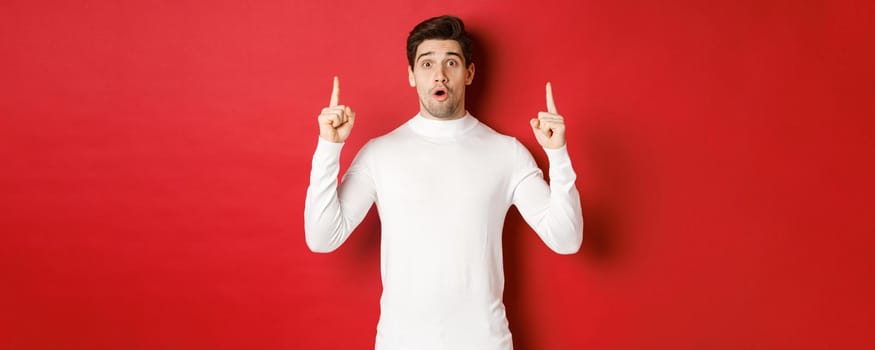 Concept of winter holidays. Good-looking young man with bristle, wearing white sweater, showing christmas advertisement on copy space, pointing fingers up and looking surprised, red background.