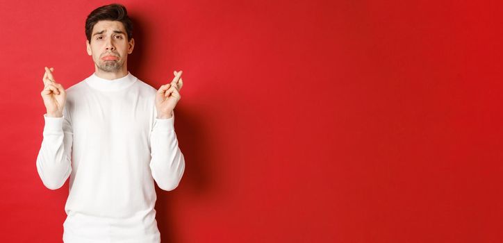 Image of sad and gloomy guy in white sweater, crying with fingers crossed, waiting for something or praying, standing over red background.