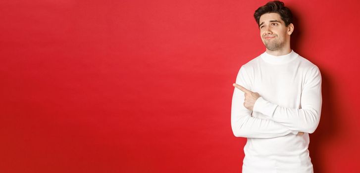 Concept of winter holidays. Image of displeased young man in white sweater, looking and pointing finger left at something average, standing skeptical over red background.