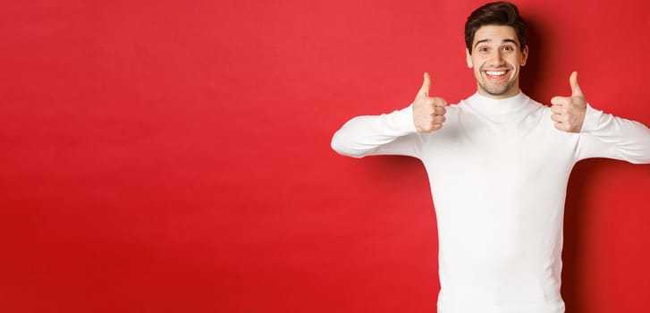 Cheerful good-looking male model in white sweater, showing thumbs-up in approval, like something good, standing over red background and smiling pleased.
