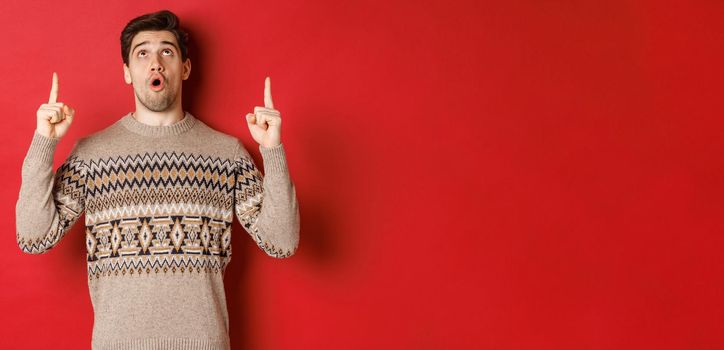 Image of amazed handsome man in christmas sweater, pointing and looking at new year promo offer, showing winter holidays advertisement on red copy space.