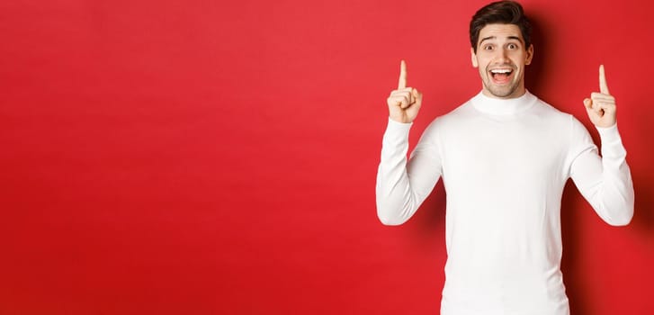 Concept of winter holidays. Good-looking young man with bristle, wearing white sweater, showing christmas advertisement on copy space, pointing fingers up and looking amazed, red background.