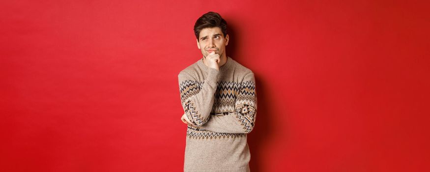 Portrait of confused man in winter sweater, thinking about christmas presents, looking troubled, searching for good gifts, standing over red background.