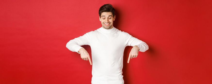 Image of dreamy good-looking man in white sweater, looking and pointing fingers down at copy space, advertisement about new year and holidays, standing over red background.