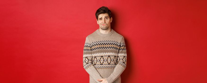 Portrait of gloomy and sad caucasian man in christmas sweater, frowning and pouting indecisive, having bad new year, standing over red background.