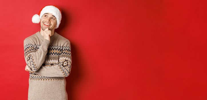 Image of happy young man in santa hat and christmas sweater, imaging something, thinking about new year gifts and smiling, looking at upper left corner, standing over red background.