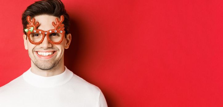 Concept of winter holidays, christmas and celebration. Close-up of attractive brunette guy in party glasses, smiling and looking happy, standing against red background.