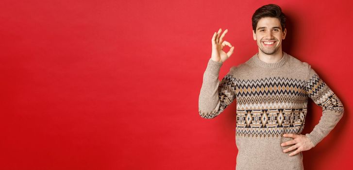 Concept of christmas celebration, winter holidays and lifestyle. Portrait of attractive man in xmas sweater, smiling happy and showing okay sign, praise something good, red background.