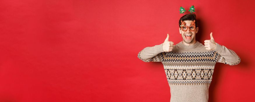 Portrait of satisfied and happy man in christmas sweater and party glasses, showing thumbs-up, wishing happy new year, standing over red background.