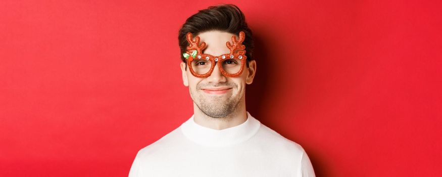 Concept of winter holidays, christmas and celebration. Close-up of attractive smiling male model in party glasses, standing against red background.