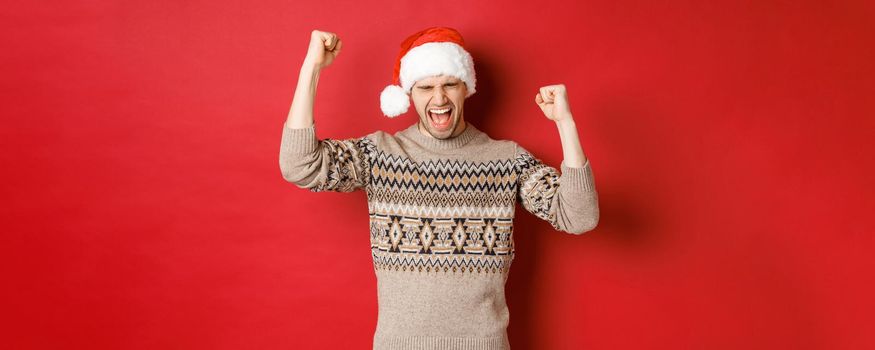 Satisfied handsome man, feeling lucky and happy, shouting for joy and making fist pumps, celebrating victory or win, receive awesome christmas gift, standing in santa hat over red background.