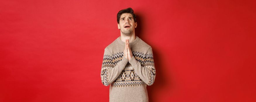 Image of nervous and hopeful man praying to God, begging for help with christmas, wearing winter sweater, looking up and pleading, standing over red background.