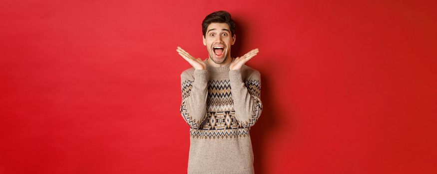 Portrait of surprised and excited guy in christmas sweater, reacting to new year present, smiling amazed, standing over red background.