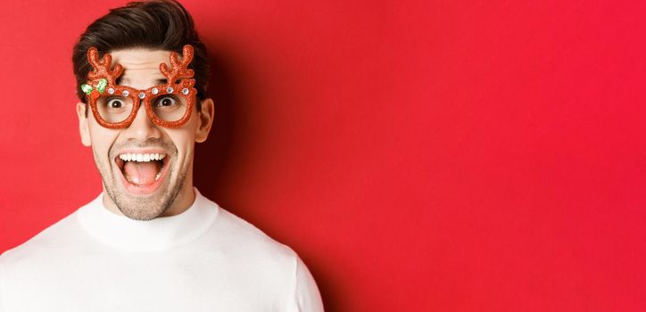 Concept of winter holidays, christmas and celebration. Close-up of attractive guy with bristle, wearing new year party glasses and looking amazed at promo offer, standing over red background.