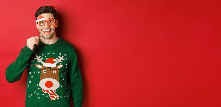 Portrait of handsome smiling man in christmas sweater and party glasses, looking surprised and happy, celebrating new year and having fun, standing against red background.