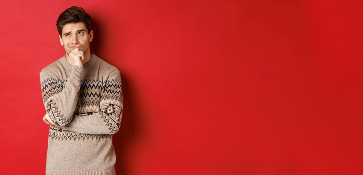 Portrait of confused man in winter sweater, thinking about christmas presents, looking troubled, searching for good gifts, standing over red background.