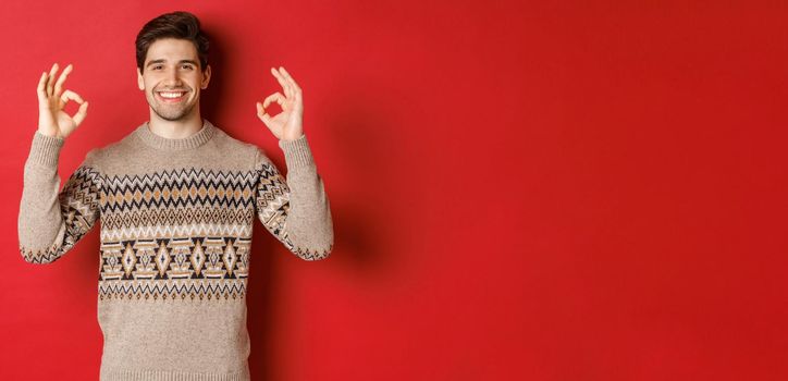 Concept of christmas celebration, winter holidays and lifestyle. Satisfied handsome man in sweater, showing okay signs and smiling pleased, recommending something, red background.