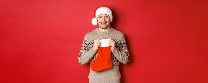 Concept of winter holidays, new year and celebration. Image of surprised and happy man receiving gift from secret santa inside christmas stocking bag, open gift and looking thankful.