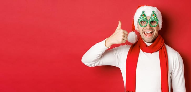 Concept of christmas, winter holidays and celebration. Cheerful guy in santa hat and party glasses, showing thumb-up and smiling pleased, standing over red background.