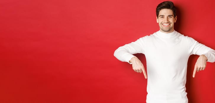 Image of smiling handsome man in white sweater inviting visit page, pointing fingers down and showing christmas banner, standing against red background.