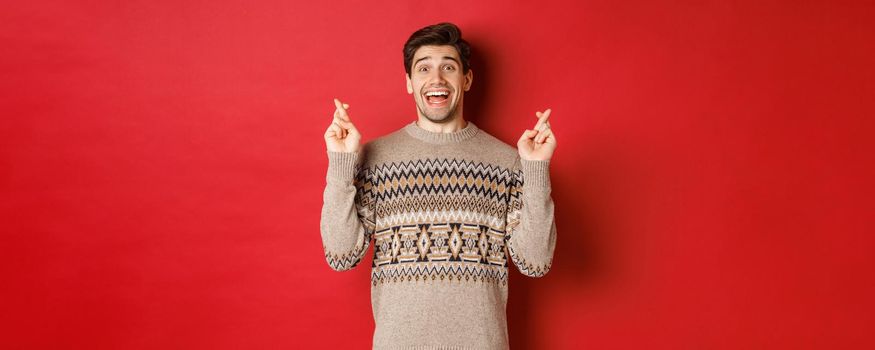 Image of excited and hopeful young man making wish, wearing winter sweater, crossing fingers for good luck and smiling, anticipating christmas eve, standing over red background.