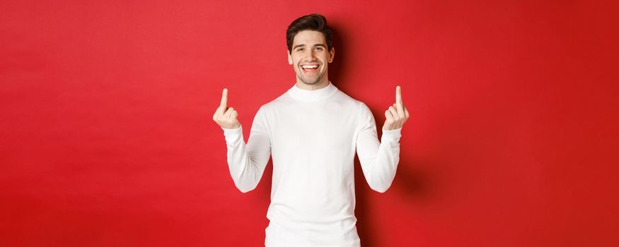 Image of rude and unbothered man laughing while showing middle-fingers, telling to fuck-off, standing over red background.