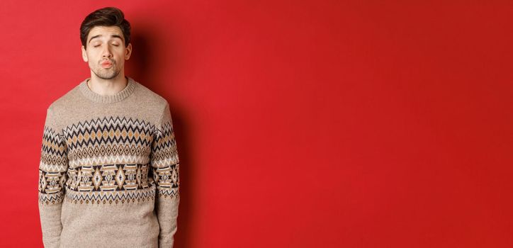 Image of handsome man in christmas sweater pucker lips and close eyes, waiting for kiss under mistletoe, standing over red background.