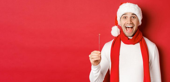 Concept of christmas, winter holidays and celebration. Close-up of happy handsome man in santa hat and scarf, having fun on new year party, holding sparkler and smiling, red background.