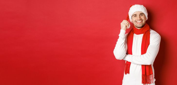 Portrait of happy man enjoying christmas holidays, wearing santa hat and scarf, smiling joyfully, standing over red background.