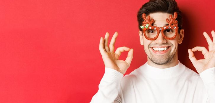 Concept of winter holidays, christmas and celebration. Close-up of attractive man in party glasses, showing okay signs and smiling, praise something good, red background.