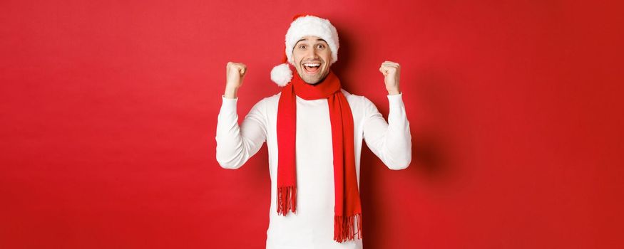 Portrait of happy and excited man in santa hat and scarf, rejoicing and winning something, celebrating new year, standing over red background.