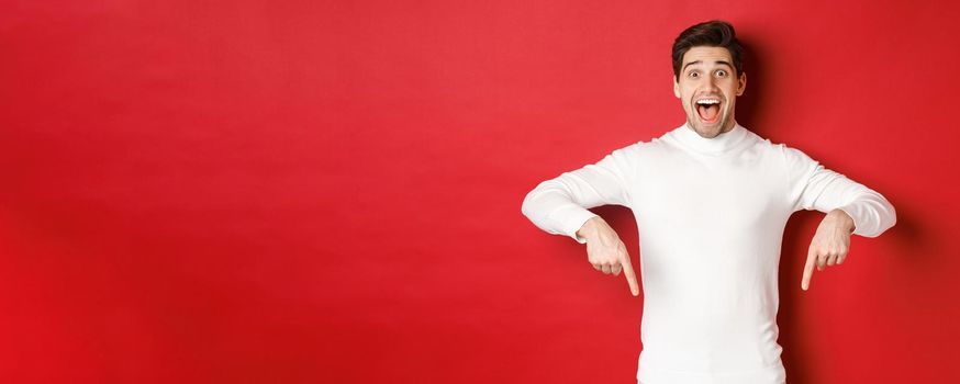 Image of amazed handsome guy in white sweater, reacting excited to christmas advertisement, pointing fingers down, showing logo, standing over red background.