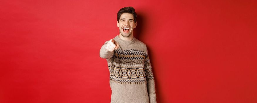 Concept of christmas celebration, winter and lifestyle. Happy attractive man in xmas sweater, wishing happy holidays and pointing at camera, standing over red background.