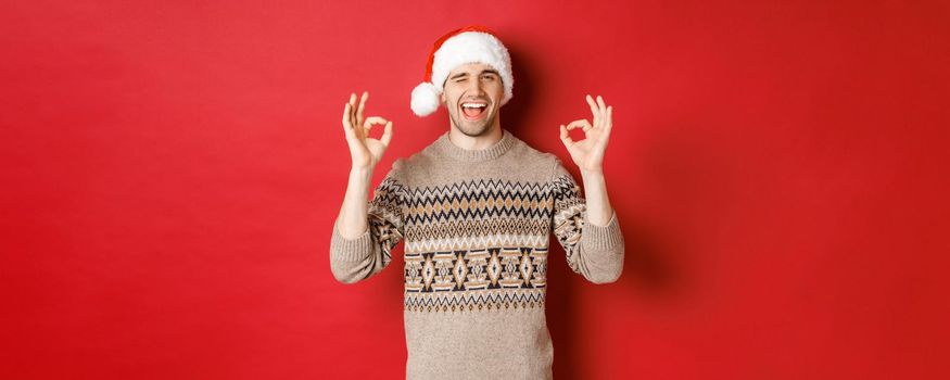 Image of joyful attractive guy in sweater and santa hat, wishing merry christmas, showing okay signs and winking at camera, celebrating new year, standing over red background.