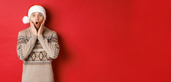 Image of amazed and speechless man in santa hat, drop jaw and staring at awesome christmas offer, standing over red background.